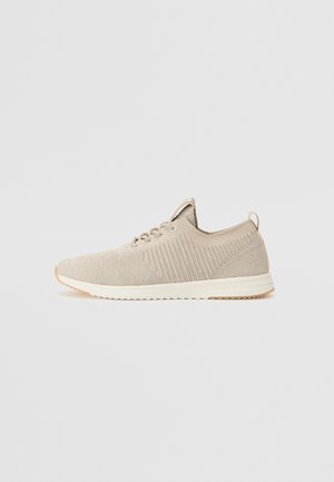 Világos bézs kötött sportcipők, texturált felsőrésszel, fehér talppal és hátsó hurokkal. Finom bordázott részletek találhatók az oldalain.