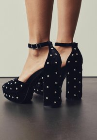 Juodos aksominės platformos kulkšnies dirželiais, papuoštos vienodais sidabro spaudais, užsidėtos ant plikų kojų prieš neutralią fono.