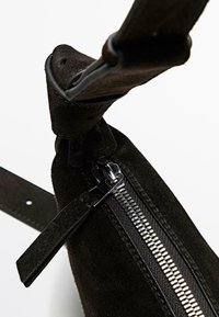 Nahaufnahme einer schwarzen Wildledertasche mit silbernem Reißverschluss und passendem Wildleder-Reißverschlussanhänger, die Textur und Details des Trägerriemens hervorhebt.