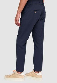 Pantaloni blu navy con vestibilità rilassata, dotati di vita elastica e una tasca posteriore singola. Indossati con sneakers color marrone chiaro e dettagli bianchi.