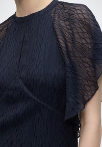 Gros plan sur une robe bleu foncé texturée avec une manche volante transparente et à motif, ainsi qu'un petit détail d'encolure en forme de trou de serrure.