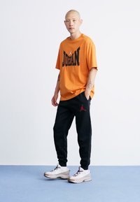 Mand står med hænderne i lommerne iført en orange Jordan T-shirt, sorte Jordan sweatpants og hvide sneakers foran en hvid baggrund.