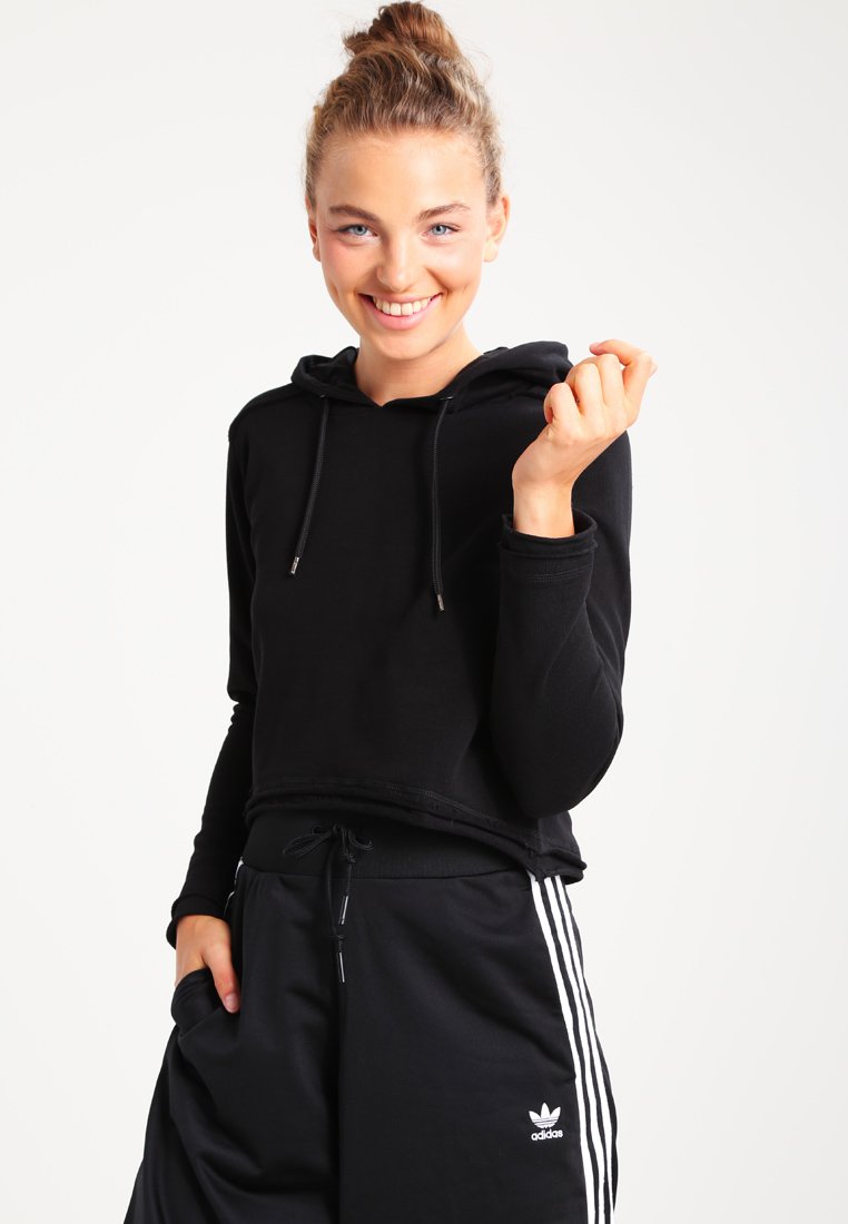 Fekete cropped kapucnis felső húzózsinórral, fekete melegítőnadrággal párosítva, amelyen fehér függőleges csíkok és Adidas logó található a lábszáron.