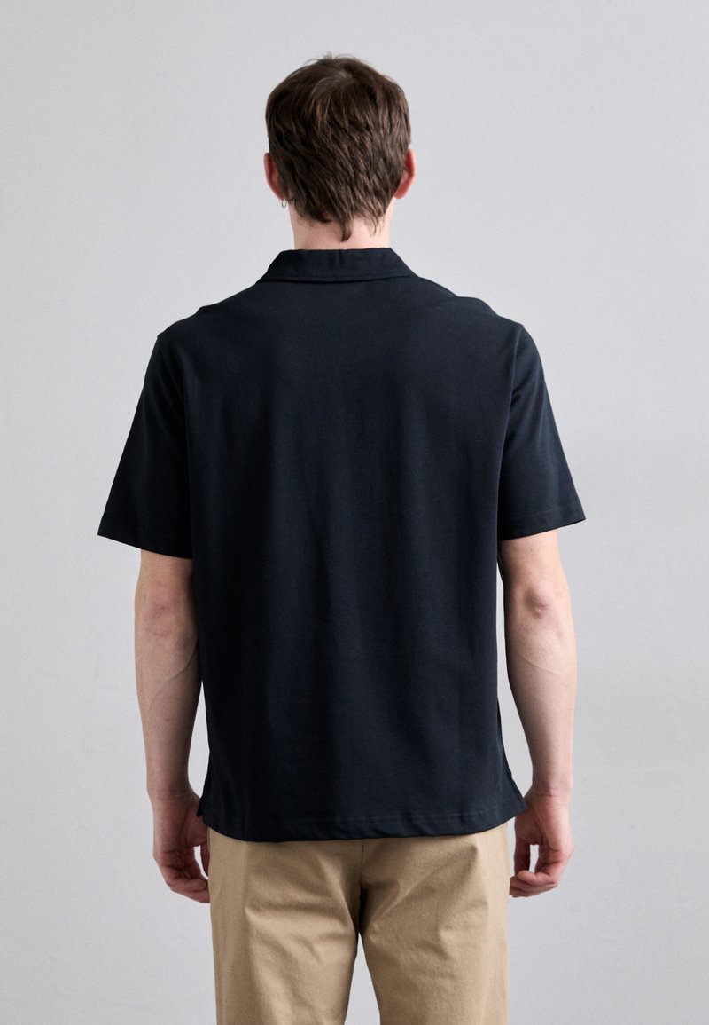 Svart kortärmad polo med klassisk krage, slät textur och rena linjer. Bärs med beiga byxor, vilket visar en enkel design.