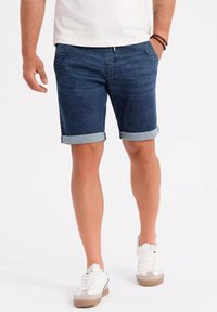 Pantalones cortos de mezclilla en azul oscuro con dobladillos enrollados, que cuentan con bolsillos laterales y cremallera. Combinados con zapatillas blancas y una camiseta ajustada.