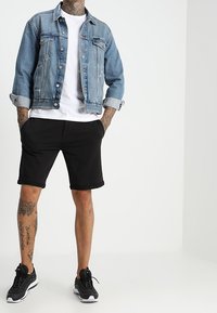 Chaqueta de mezclilla, azul claro con bolsillos de botón, sobre una camiseta blanca. Pantalones cortos negros, hasta la rodilla. Zapatillas deportivas negras. Tatuajes visibles en las piernas.