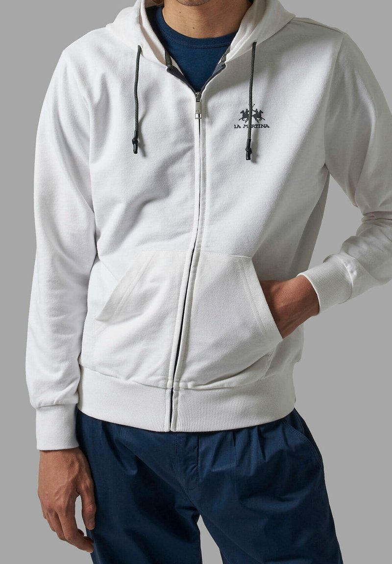 Homme portant un sweat à capuche blanc zippé avec poches avant et cordons noirs, associé à un pantalon bleu foncé sur fond gris.