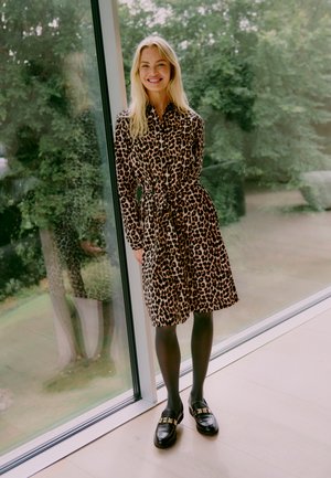 Robe imprimée léopard avec des manches longues, taille ceinturée et ourlet au genou. Des collants noirs et des mocassins épais complètent la tenue.