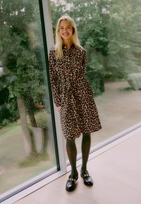 Obleka z leopardjim odtisom z dolgimi rokavi, pasom in dolžino do kolen. Črne najlonke in debele loafers čevlji dopolnjujejo obleko.