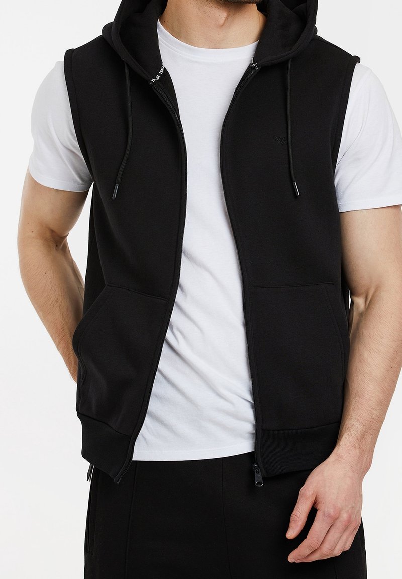 Veste sans manches noire avec capuche zippée à l'avant, deux poches frontales, cordon de serrage à la capuche et texture douce, portée avec un t-shirt blanc.