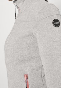 Hellgraue Fleecejacke mit hohem Kragen, Seitentaschen mit rosa Reißverschlüssen und einem schwarzen Badge auf dem linken Ärmel.