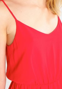 Czerwona sukienka bez rękawów, wykonana z lekkiego materiału. Posiada dekolt w kształcie litery V oraz elastyczny pasek w talii, co zapewnia luźne dopasowanie. Gładka faktura, prosty design.