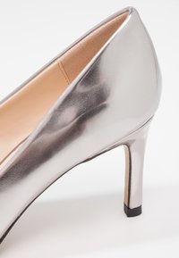 Escarpin en cuir verni argenté à talon haut, avec un bout pointu et une silhouette effilée, et un détail de talon noir contrastant. Texture lisse et réfléchissante.