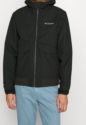 Schwarze Columbia-Kapuzenjacke mit Frontreißverschluss, Klappentaschen und verstellbaren Manschetten, getragen von einer Person in hellblauen Hosen.