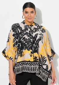 Blouse à imprimé floral avec des motifs noirs et jaunes sur un fond beige clair, dotée d'un devant boutonné et d'un ourlet asymétrique.
