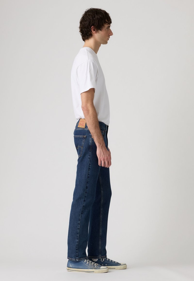 Giovane uomo in piedi di profilo che indossa una maglietta bianca, jeans blu e sneakers blu su uno sfondo chiaro uniforme.