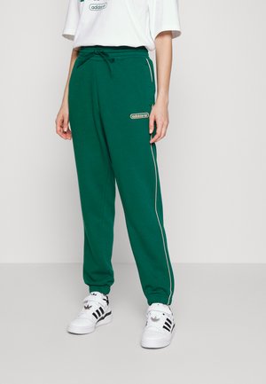 Pantalones de chándal verdes con cintura ajustable, que presentan una franja lateral de color crema y un logo bordado en el muslo. Combinados con zapatillas blancas.
