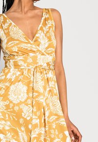 Robe portefeuille florale en tissu jaune moutarde avec des motifs botaniques blancs. Dispose d'un décolleté en V profond et d'une ceinture à nouer à la taille.