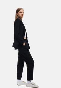 Zwarte op maat gemaakte blazer over een witte top, gecombineerd met zwarte afgebouwde broeken en witte schoenen. Gladde stof, gestructureerd ontwerp en casual stijl.