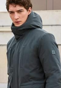 Jachetă puf gri închis cu guler înalt și glugă. Dispune de două buzunare frontale și un patch mic cu marcaj pe mâneca stângă. Textură netedă.