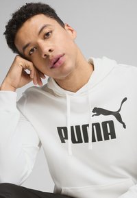 Hanorac alb cu un logo "PUMA" negru și o siluetă de pisică. Dispune de șnururi, o textură moale și o croială lejeră.