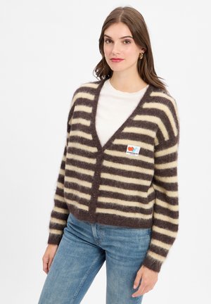 Frau in einem braun-cream gestreiften, flauschigen Cardigan über einem weißen Oberteil und blauen Jeans, vor einem schlichten weißen Hintergrund stehend.