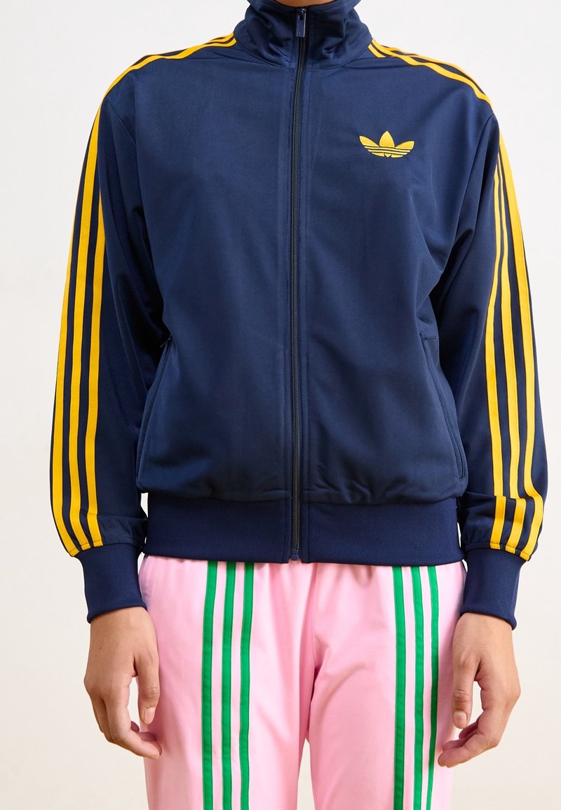 Veste de sport zippée marine avec des rayures jaunes et un logo ; présente un col haut et des poignets élastiques. Associée à un pantalon rose avec des rayures vertes.