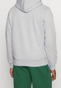 Uomo che indossa un hoodie grigio chiaro e pantaloni verdi, visto da dietro, in piedi contro uno sfondo chiaro e semplice.