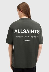 Camiseta de algodón verde oscuro con mangas cortas y cuello redondo. Presenta un gran texto blanco en la parte posterior: "ALLSAINTS" y "LONDRES".