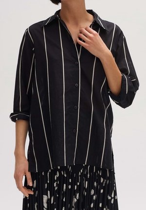 Personne portant une chemise noire à manches longues avec des rayures verticales blanches et une jupe noire plissée avec des taches abstraites blanches.