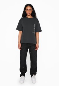 Grå oversized t-shirt med korte ærmer, prydet med et minimalistisk hvidt grafik på venstre side, parret med mørke jeans og hvide sneakers.