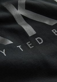 Schwarzer Stoff mit einer glatten Textur, auf dem in fetten silbernen Buchstaben "TED B." steht. Das Design enthält markante geometrische Akzente.