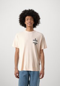 Light pink cotton t-shirt with short sleeves, featuring a graphic design in black and gray on the upper left chest. Casual fit.