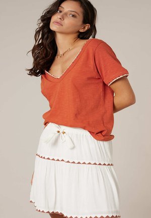 Femme aux cheveux bruns ondulés portant un haut orange à manches courtes avec bordure et une jupe blanche avec taille à cordon et coutures décoratives marron.