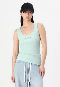 Débardeur côtelé vert menthe avec le texte "CAP" en lettres brillantes, associé à un jean baggy bleu clair et une casquette de baseball noire.