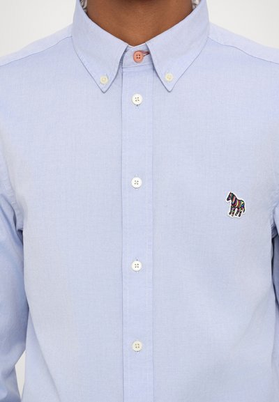 Chemise en coton bleu clair à manches longues, ornée d'une petite broderie de zèbre coloré sur la poitrine gauche et de boutons multicolores.