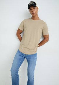 T-shirt en coton beige avec un col rond, associé à un jean ajusté bleu clair. La personne porte une casquette noire avec un logo.