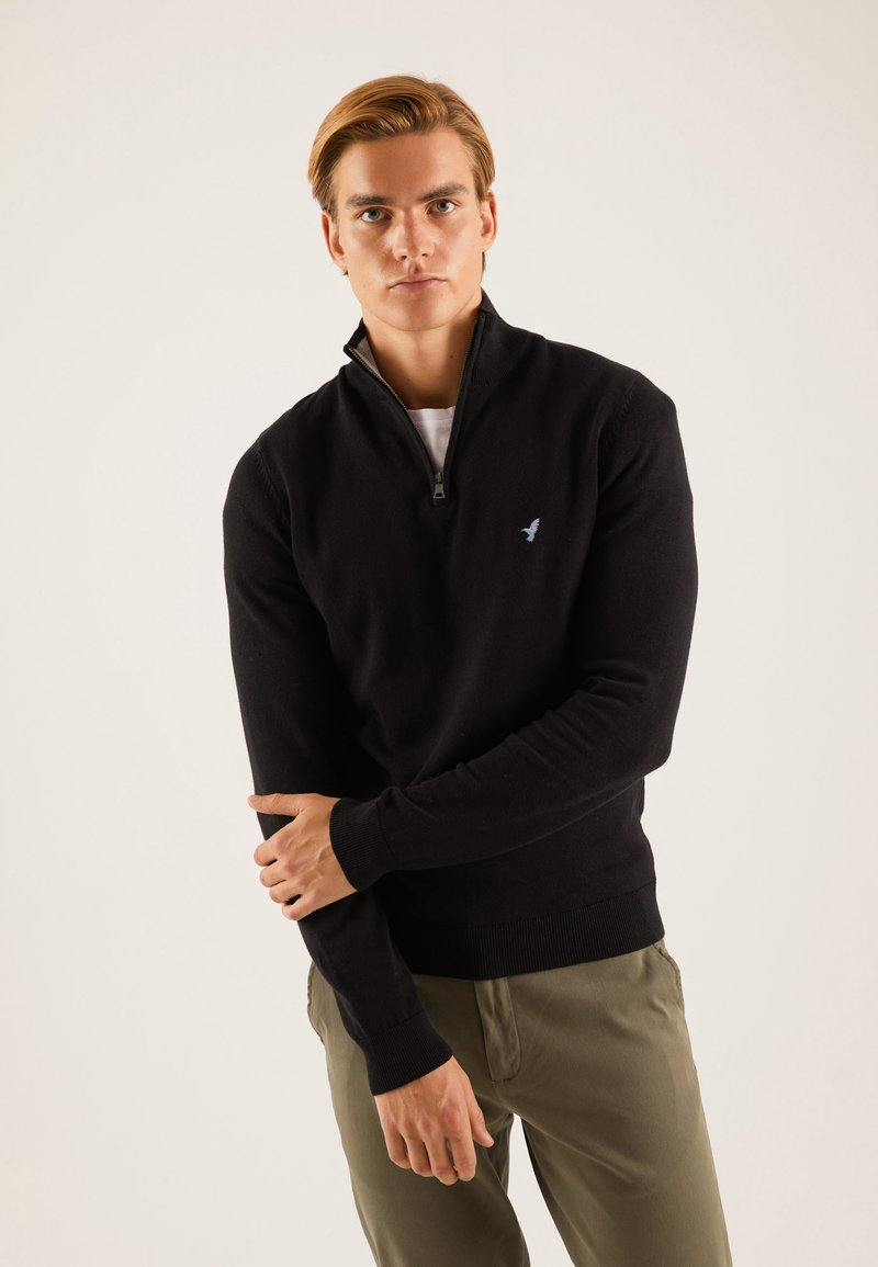 Sweat-shirt zippé noir avec un ourlet et des poignets côtelés. Présente un petit logo bleu sur la poitrine. Porté par-dessus une chemise blanche.