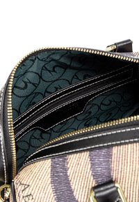 Väskan har en mönstrad utsida i beige, accentuerad med mörklila och svart. Inuti finns ett grönt tyg med en upprepad logotyp samt en dragkedjeficka.