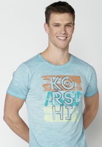 Světle modré tričko z měkké tkaniny má grafický potisk v několika barvách, včetně oranžové a zelené, s stylizovaným textem a detaily palmy.