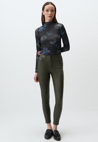 Zwarte geribbelde lange mouwen top met bloemenpatroon, gecombineerd met olijfgroene geplooide broek. Model staat en toont een slanke silhouet en eenvoudige zwarte schoenen.