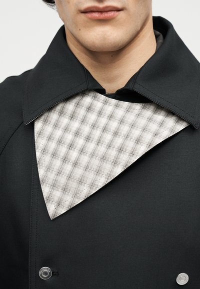Άντρας με μαύρο παλτό με φαρδιά γιακά και ορατό λευκό και γκρι καρό εσωτερικό στρώμα κάτω από τον γιακά.