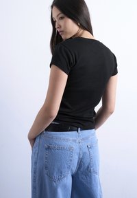 Νεαρή γυναίκα με μακριά σκουρόχρωμα μαλλιά, φορώντας εφαρμοστό μαύρο t-shirt και μπλε τζιν, όρθια με τα χέρια στις τσέπες, στραμμένη ελαφρώς προς τα πλάγια.