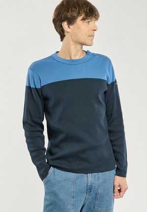 Homme portant un pull à manches longues color block avec des épaules bleues et un corps bleu foncé, associé à un jean bleu clair, regardant sur le côté.