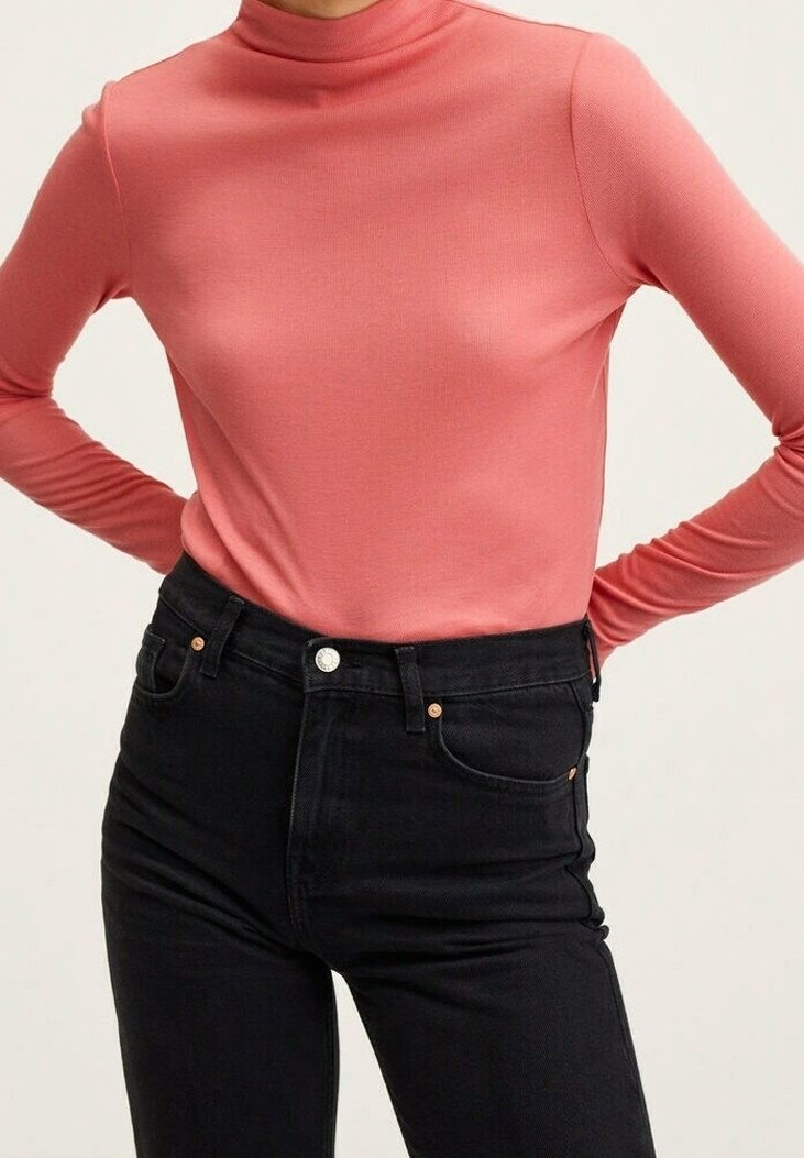 Une personne portant un col roulé corail à manches longues rentré dans un jean noir taille haute, les mains sur les hanches, contre un fond neutre.