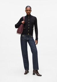 Cardigan nero a coste con bottoni dorati, abbinato a jeans di jeans scuri con tasche frontali e una borsa bordeaux. Stivali marroni con tacco.