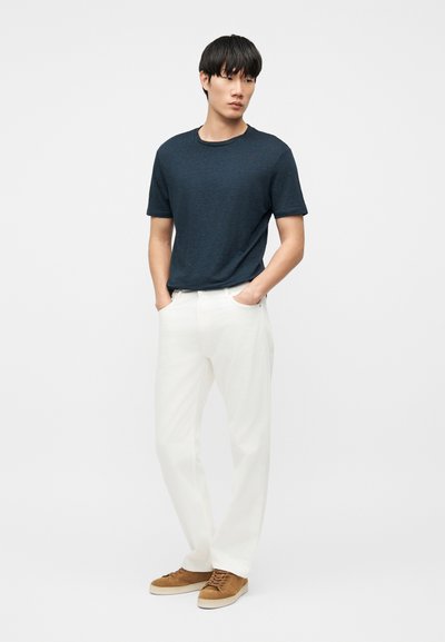 Νεαρός άνδρας στέκεται με τα χέρια στις τσέπες, φοράει μπλε σκούρο μπλουζάκι, λευκό παντελόνι και καφέ casual παπούτσια μπροστά σε απλό φόντο.