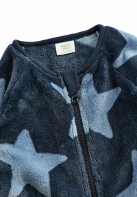 Tummansininen fleece-vaate, jossa on vaaleansinisiä tähtikuvioita, varustettuna vetoketjulla ja ribbaamalla kaulalla. Pehmeä rakenne, rento muotoilu.