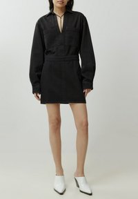 Zwarte denim jurk met lange mouwen, knoopsluiting aan de voorkant, twee borstzakken, een taille met trekkoord, in combinatie met witte, hooggehakte muiltjes. Glad textuur.