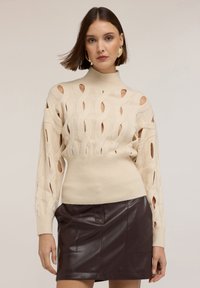 Beżowy sweter dzianinowy z wycięciami, wysoka szyja, ściągacze i dopasowana talia, w połączeniu z ciemnobrązową skórzaną mini spódnicą.